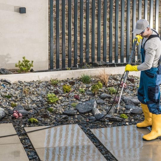 Caucasian Middle Aged Worker Washing Concrete Tiles in the Yard with a Pressure Washer Machine. House Surrounding Seasonal Maintenance.